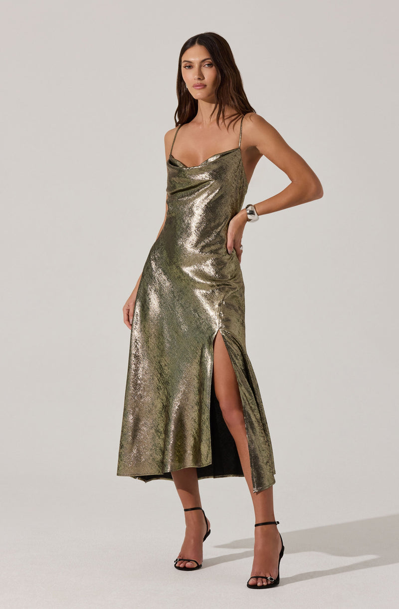 A woman is wearing a metallic, shiny, gold dress with spaghetti straps and a cowl neckline. The dress has a high slit on one side, revealing part of her leg. She is also wearing black high-heeled sandals with ankle straps.