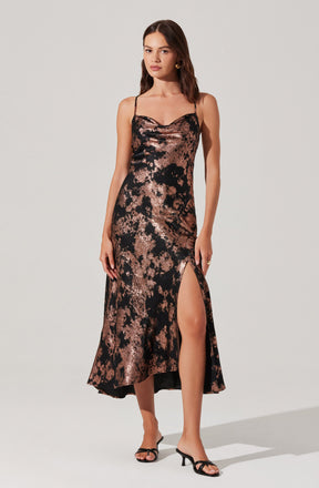A woman is wearing a sleeveless, full-length slip dress with thin spaghetti straps. The dress has a metallic, abstract marbled pattern in shades of black and rose gold, with a high slit on one side that reaches above the knee. She is also wearing black high-heeled sandals with thin straps.