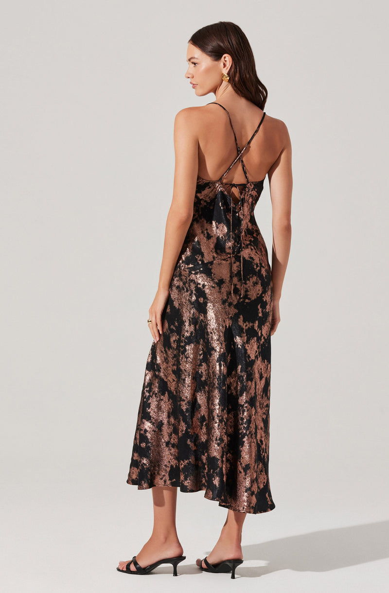 A woman is wearing a sleeveless, full-length dress with a shiny, metallic finish. The dress features a black and bronze abstract pattern, thin spaghetti straps, and a low back with a tied bow. She is also wearing black high-heeled sandals and gold earrings.