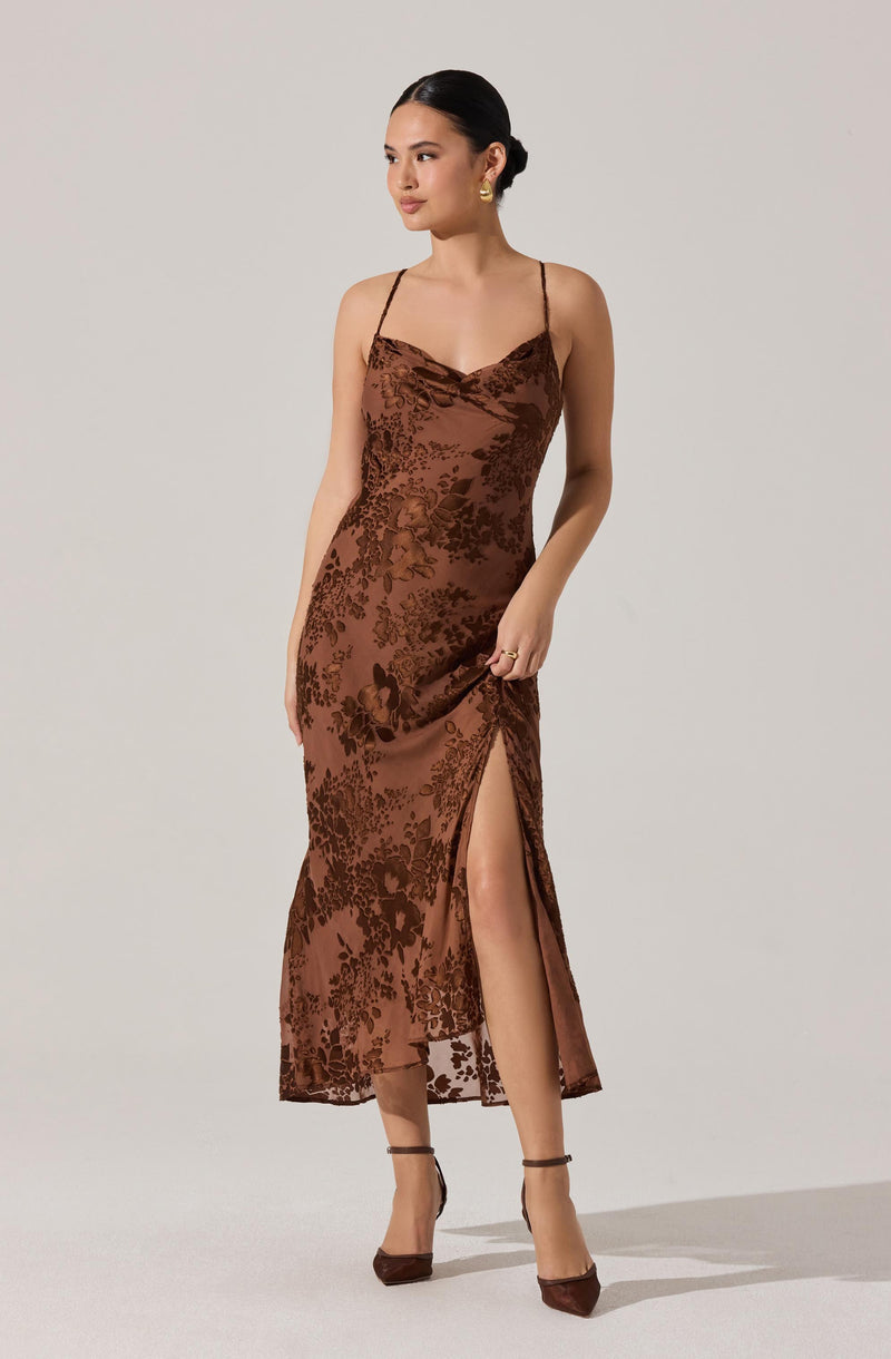 A woman is wearing a brown satin slip dress with a floral jacquard pattern. The dress features thin spaghetti straps, a cowl neckline, and a high slit up one side. She has paired the dress with matching brown high-heeled shoes.
