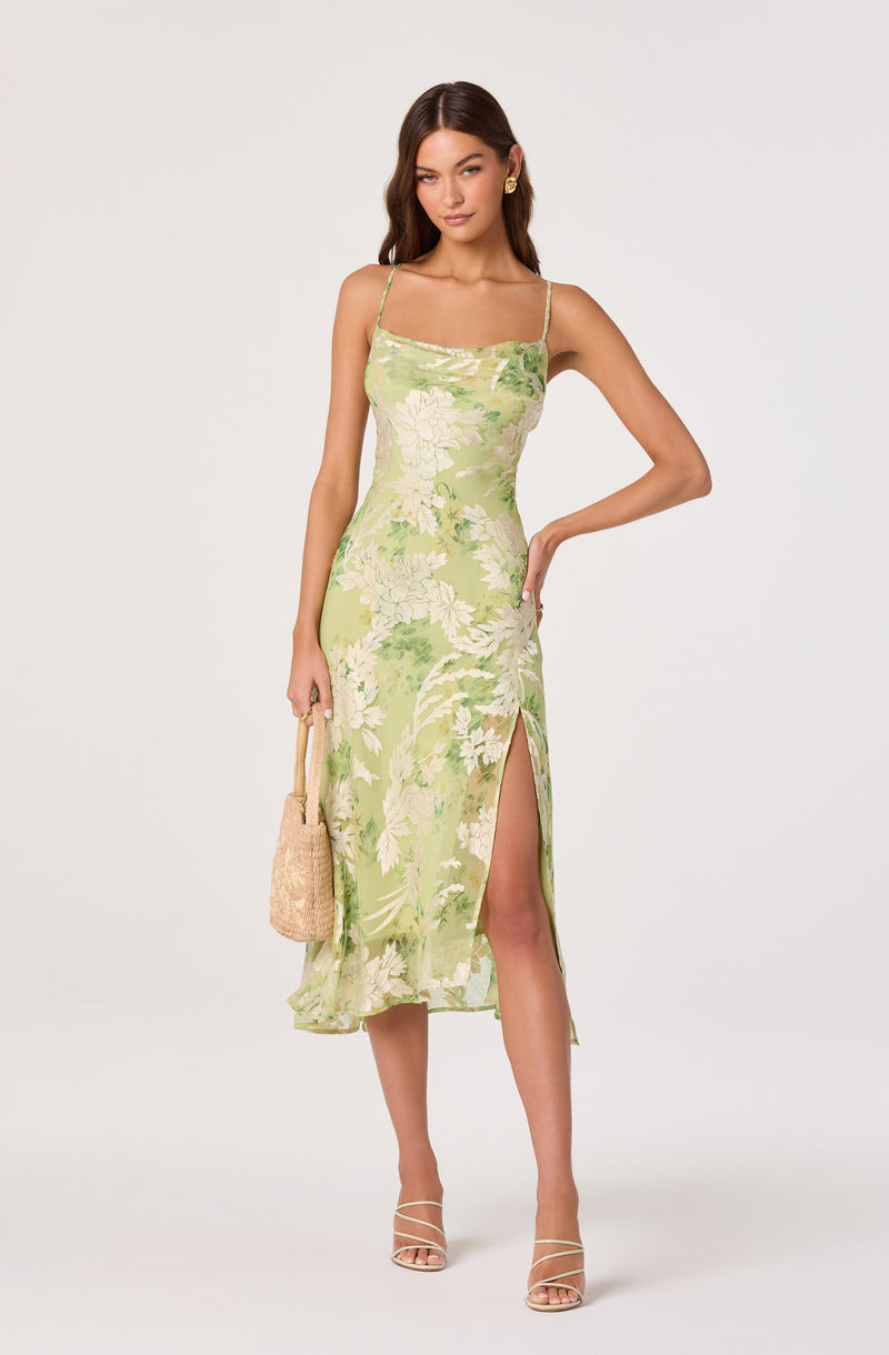 Woman wearing a green floral spaghetti strap midi dress with a slit, holding a woven bag.