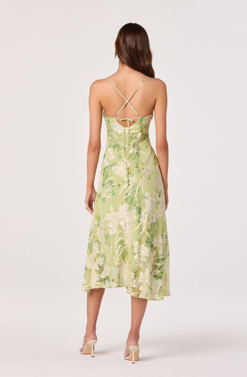 Back view of a woman in a green floral midi dress with thin crisscross straps and a bow tie.