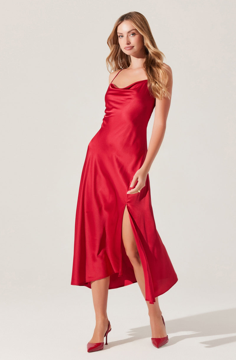 A woman is wearing a long, silky red dress with thin shoulder straps. The dress features a subtle cowl neckline, a thigh-high slit on one side, and matching red heels.