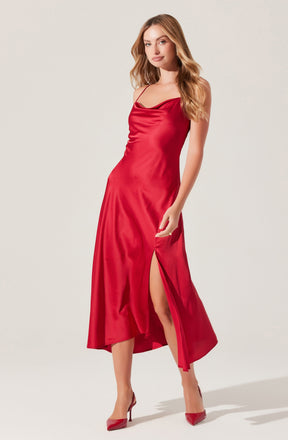 A woman is wearing a long, silky red dress with thin shoulder straps. The dress features a subtle cowl neckline, a thigh-high slit on one side, and matching red heels.