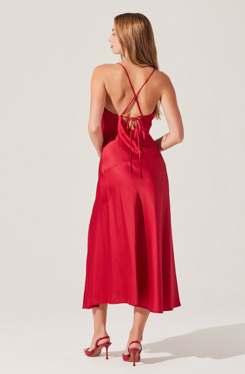 A woman is wearing a long, flowing red satin dress with spaghetti straps that crisscross at the back and tie at the waist. She is also wearing matching red high-heeled sandals.
