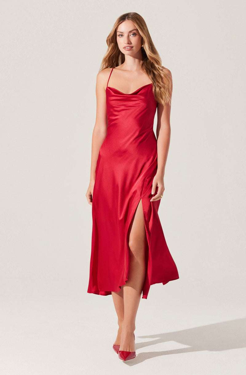 A woman is wearing a long, shiny red satin dress with spaghetti straps. The dress has a draped neckline and a high slit on one side, revealing her leg. She is also wearing matching red high heels.