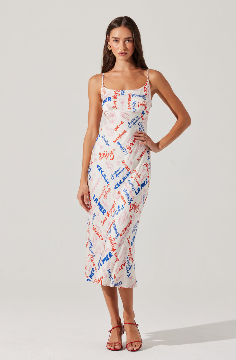 A woman is wearing a sleeveless, ankle-length dress with thin spaghetti straps. The dress is predominantly white with a colorful, abstract print consisting of various words and phrases in red and blue. She is also wearing red high-heeled sandals.