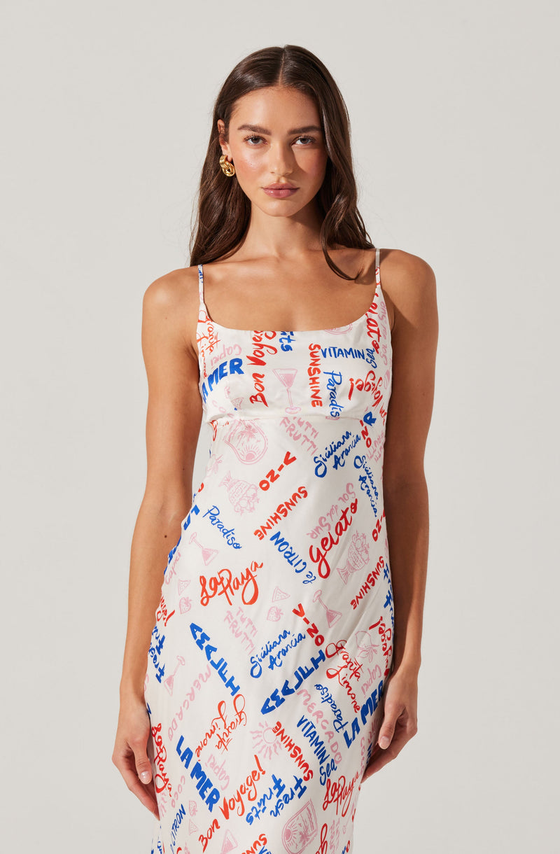A woman is wearing a sleeveless, spaghetti strap dress with a white background. The dress is decorated with various colorful text and graphic elements in red and blue, including words like "Gelato," "Summer," and "Vitamin." The overall style of the dress is casual and summery.