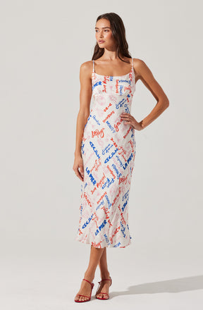 A woman is wearing a sleeveless, spaghetti strap midi dress with a white background and a colorful print of various words and phrases in red and blue. She has paired the dress with red high-heeled sandals.