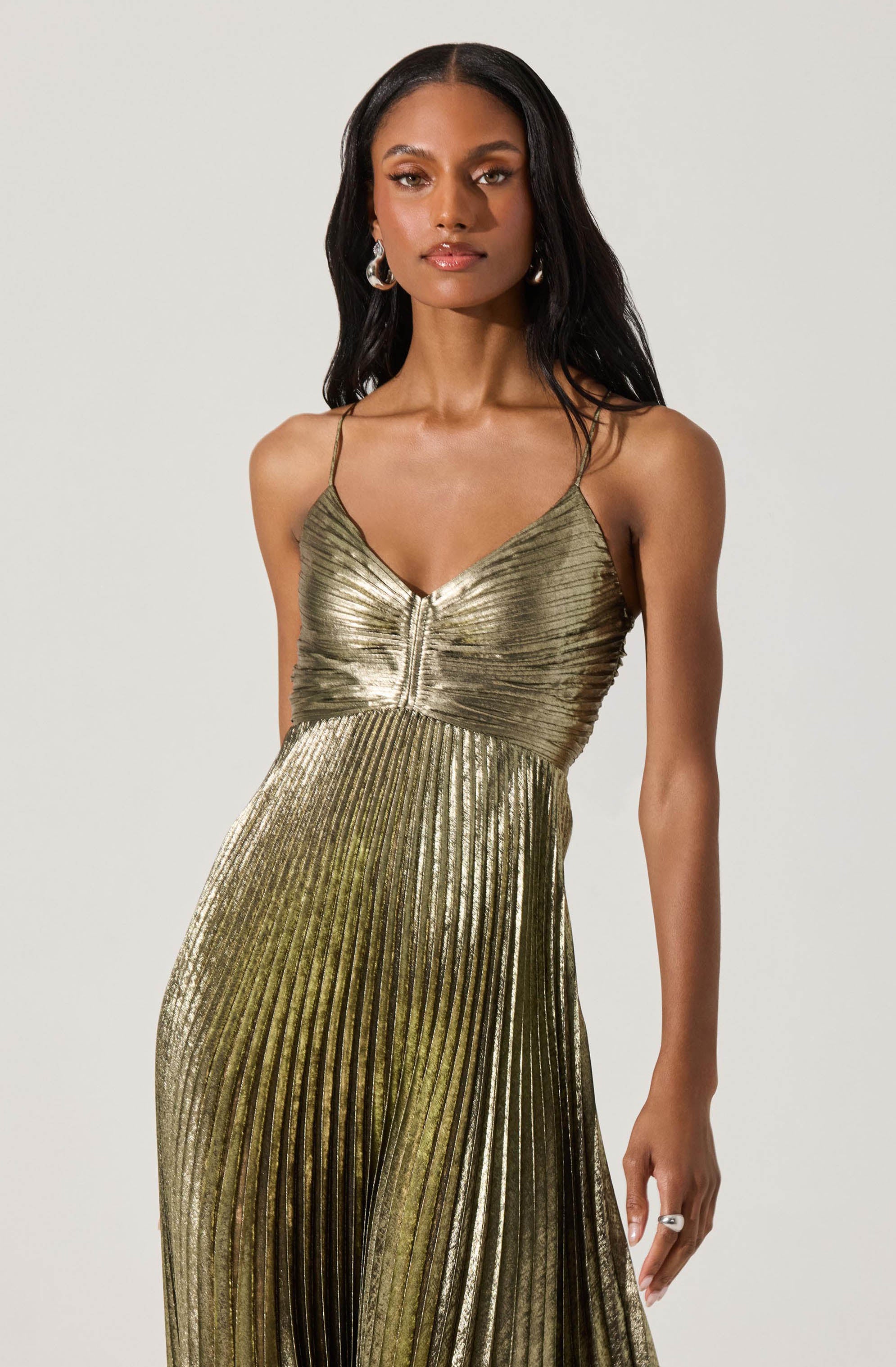 A woman is wearing a metallic gold dress with thin spaghetti straps. The dress features a fitted bodice with a gathered detail at the center and a pleated skirt that extends to the floor.