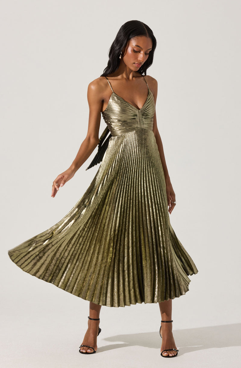 A woman is wearing a sleeveless, metallic gold dress with thin spaghetti straps. The dress features a fitted bodice with a gathered detail at the center and a flowing, pleated skirt that reaches mid-calf length. She pairs the dress with black high-heeled sandals.