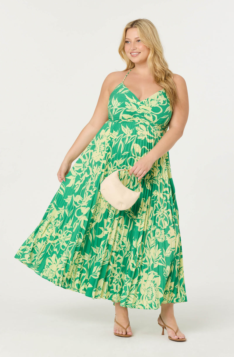 Woman in a green and yellow floral maxi dress with spaghetti straps, holding a beige handbag.