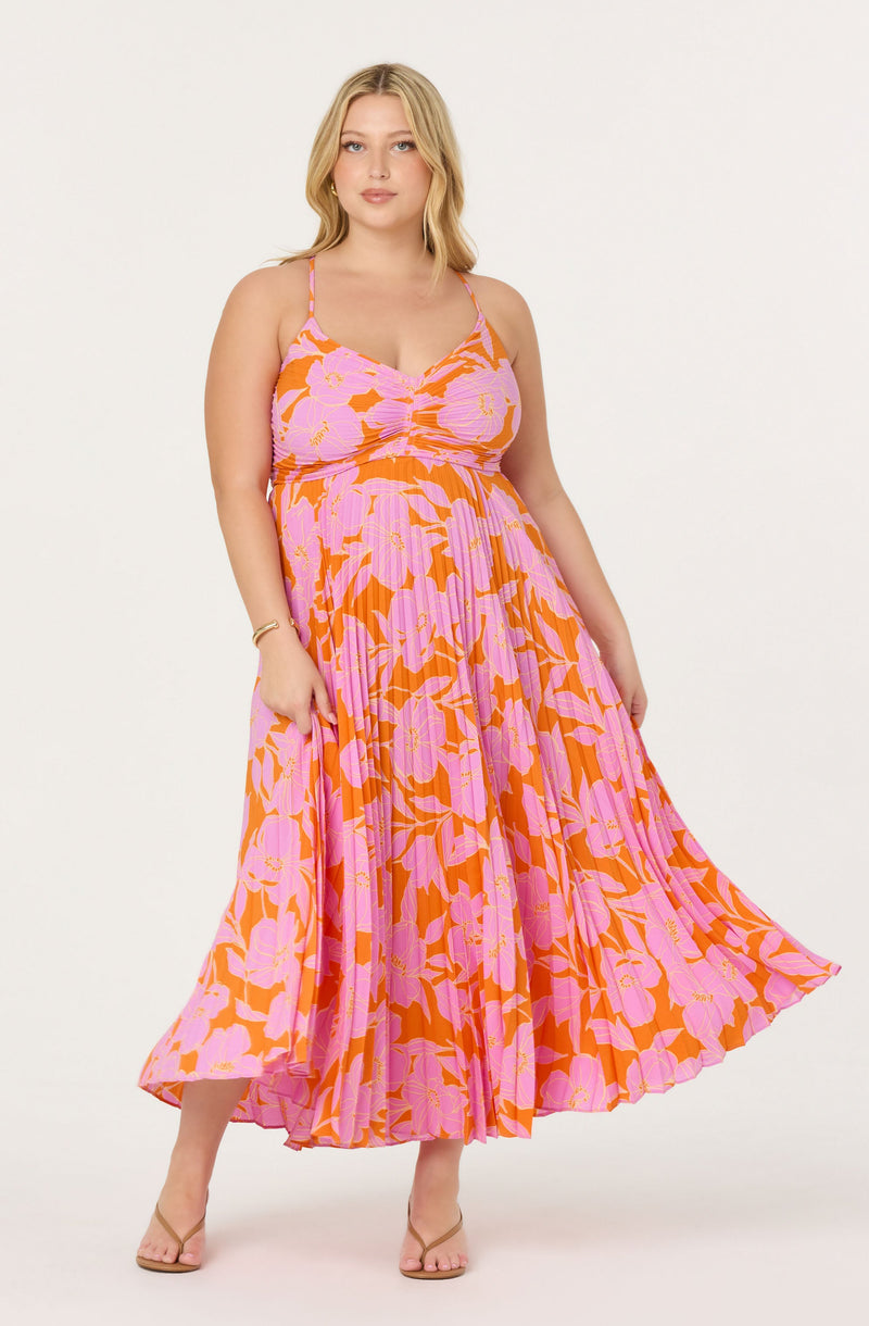 A woman is wearing a sleeveless, spaghetti strap maxi dress with a vibrant floral pattern. The dress features large pink flowers with yellow accents on an orange background, and has a ruched bodice and a pleated, flowy skirt. She is also wearing nude open-toe heels and a gold bracelet on her right wrist.