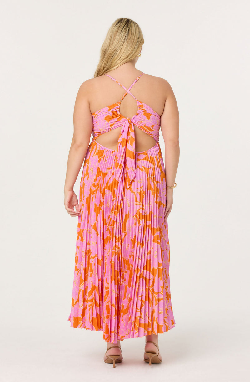 A woman is wearing a sleeveless, floor-length dress with a pink and orange floral pattern. The dress features crisscrossing spaghetti straps, a circular cutout in the back, and a tie at the waist. The fabric is pleated, giving it a flowing appearance. She is also wearing beige high-heeled sandals.