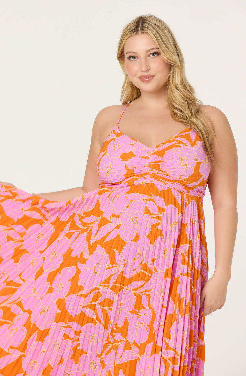 A woman is wearing a sleeveless maxi dress with spaghetti straps. The dress features a vibrant floral pattern with large pink flowers and green leaves on an orange background. The fabric has a pleated texture, and the top part of the dress is gathered and ruched at the center.