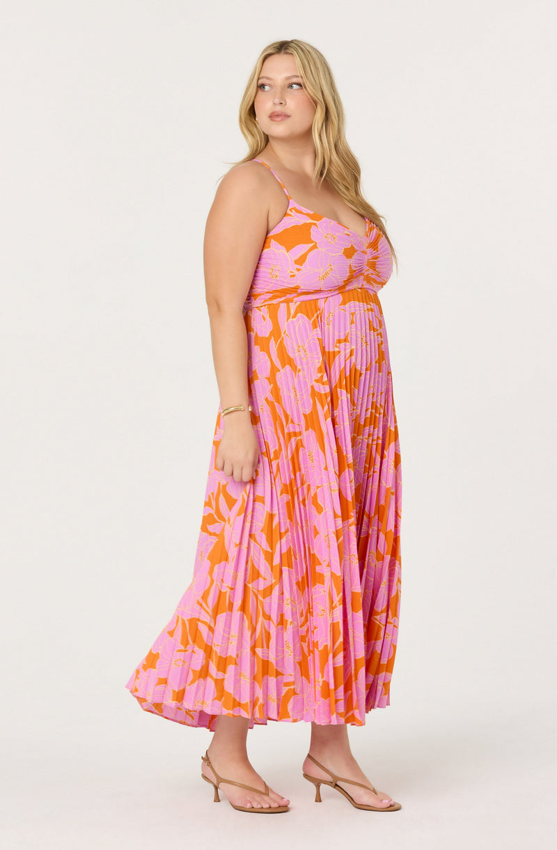 A woman is wearing a sleeveless, long, flowing dress with a vibrant floral pattern in shades of pink and orange. The dress has thin spaghetti straps and a fitted bodice that transitions into a pleated skirt. She is paired with beige, open-toed, heels with a thin strap.
