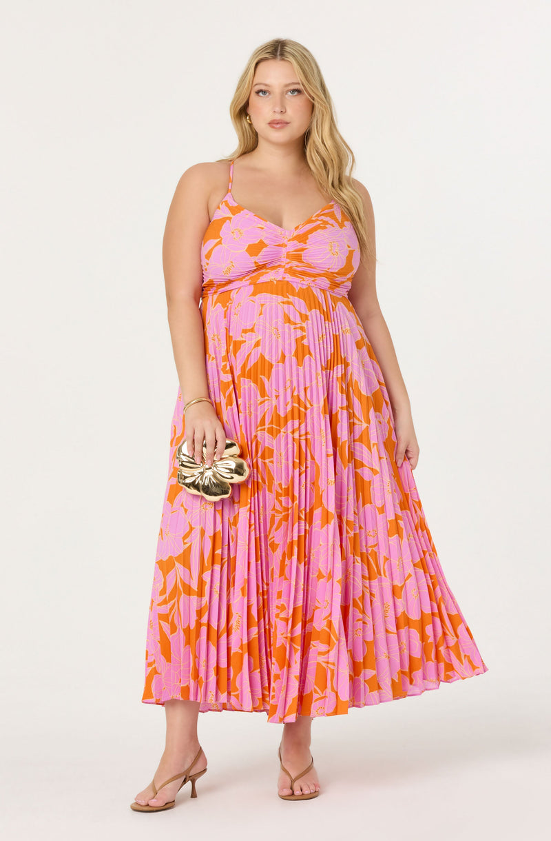 A woman is wearing a long, sleeveless, floral pleated maxi dress in shades of pink and orange. The dress features thin spaghetti straps and a gathered detail at the bust. She is holding a shiny metallic gold clutch bag with a sculptural, abstract shape. She is also wearing nude open-toed heels and gold jewelry, including earrings and a bracelet.