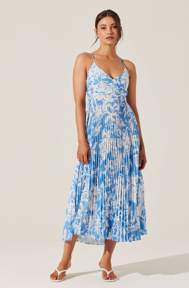 A woman is wearing a sleeveless, flowy maxi dress with thin spaghetti straps. The dress features a blue and white abstract floral pattern with pleats running vertically from top to bottom. She is also wearing white strappy high-heeled sandals.