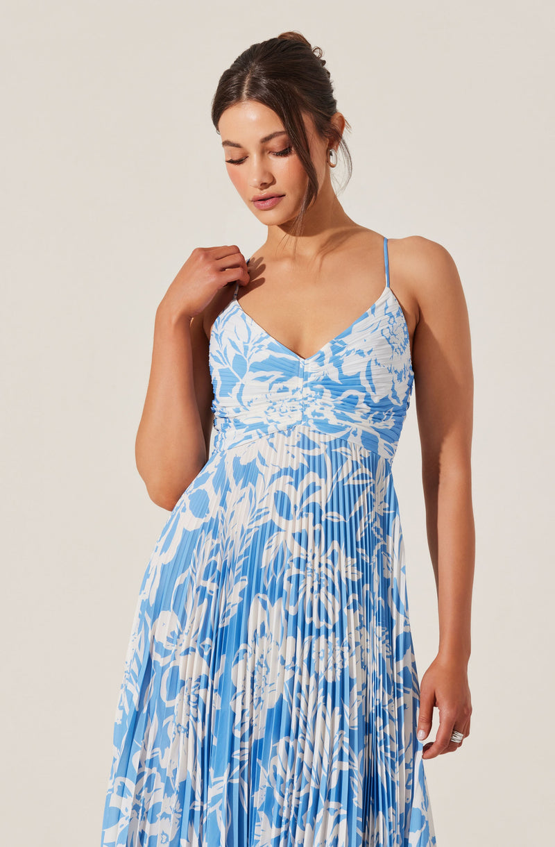 A woman is wearing a sleeveless dress with thin spaghetti straps. The dress features a ruched bodice and a flowing, pleated skirt with an abstract blue and white floral pattern.