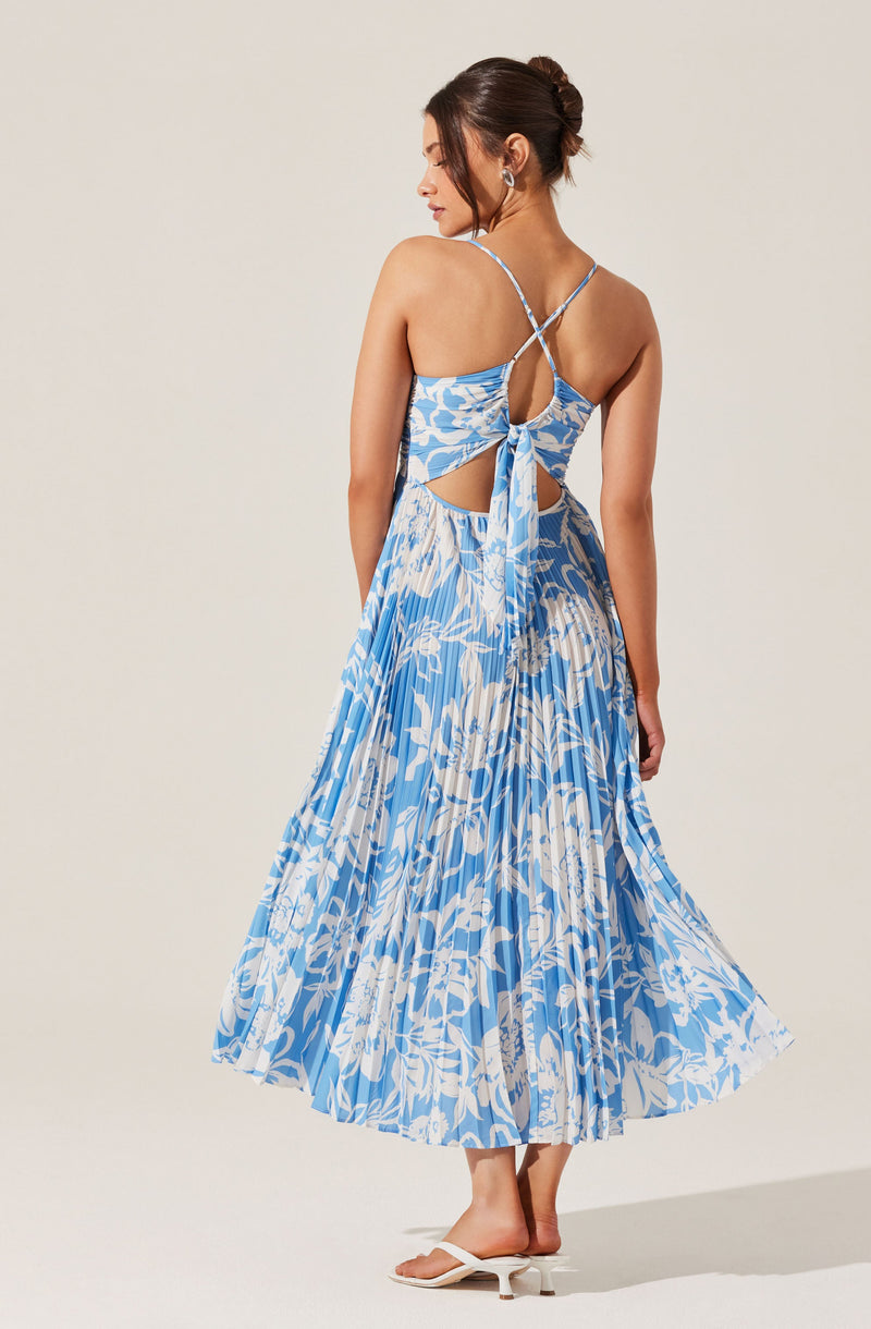 A woman is wearing a blue and white floral dress with a pleated skirt. The dress features thin, crisscross spaghetti straps on the back and cut-outs on the sides, with a tie detail at the back. She is also wearing white heeled sandals with a small heel. Her hair is styled in an elegant bun, and she has large earrings.