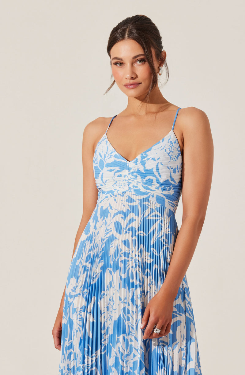 A woman is wearing a sleeveless dress with thin spaghetti straps. The dress features a fitted bodice with gathered fabric at the center and a flowing, pleated skirt. The fabric has a blue and white floral pattern.