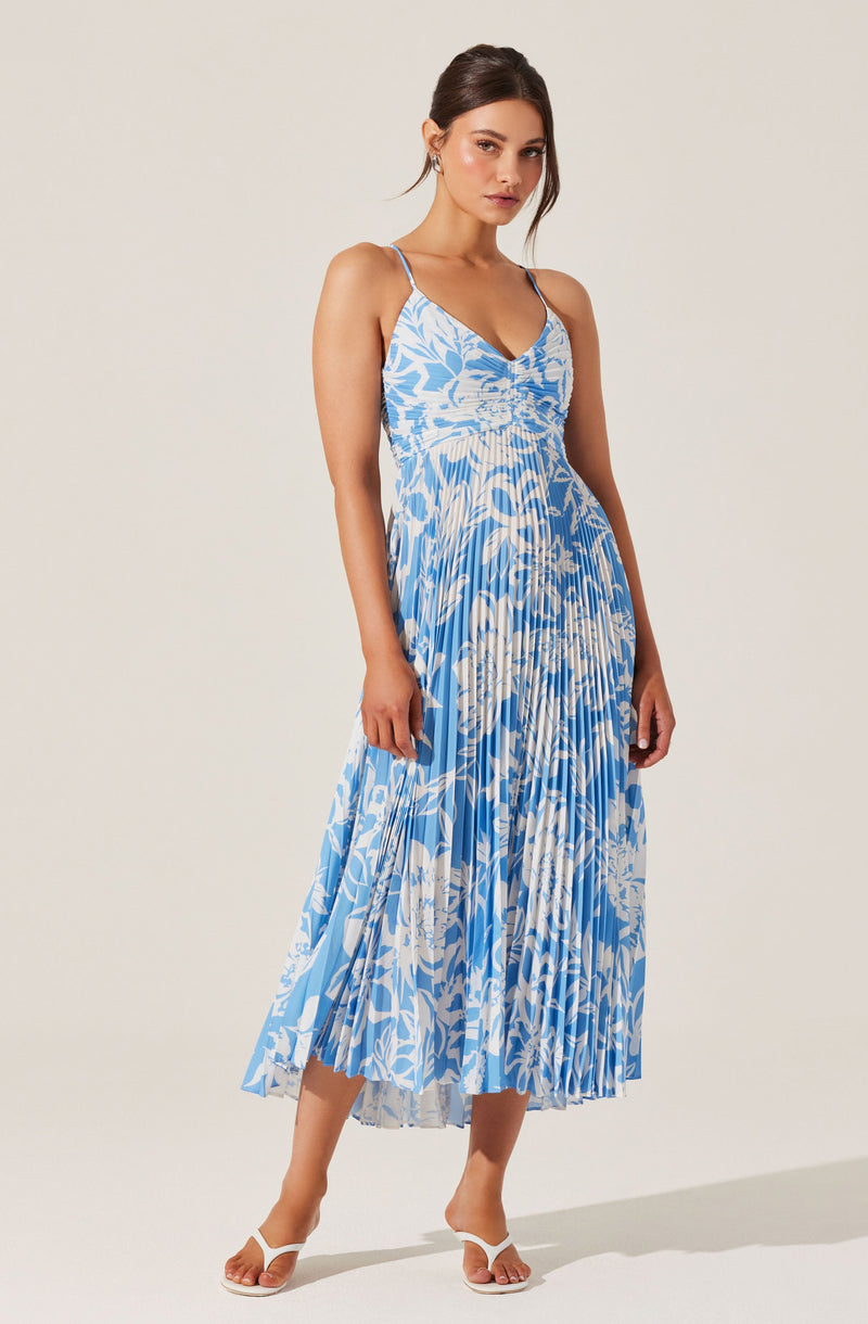 A woman is wearing a sleeveless, V-neck dress with thin spaghetti straps. The dress features a blue and white floral and leaf print, with a fitted waist and a pleated, midi-length skirt that flows down to her ankles. She is also wearing white high-heeled sandals.