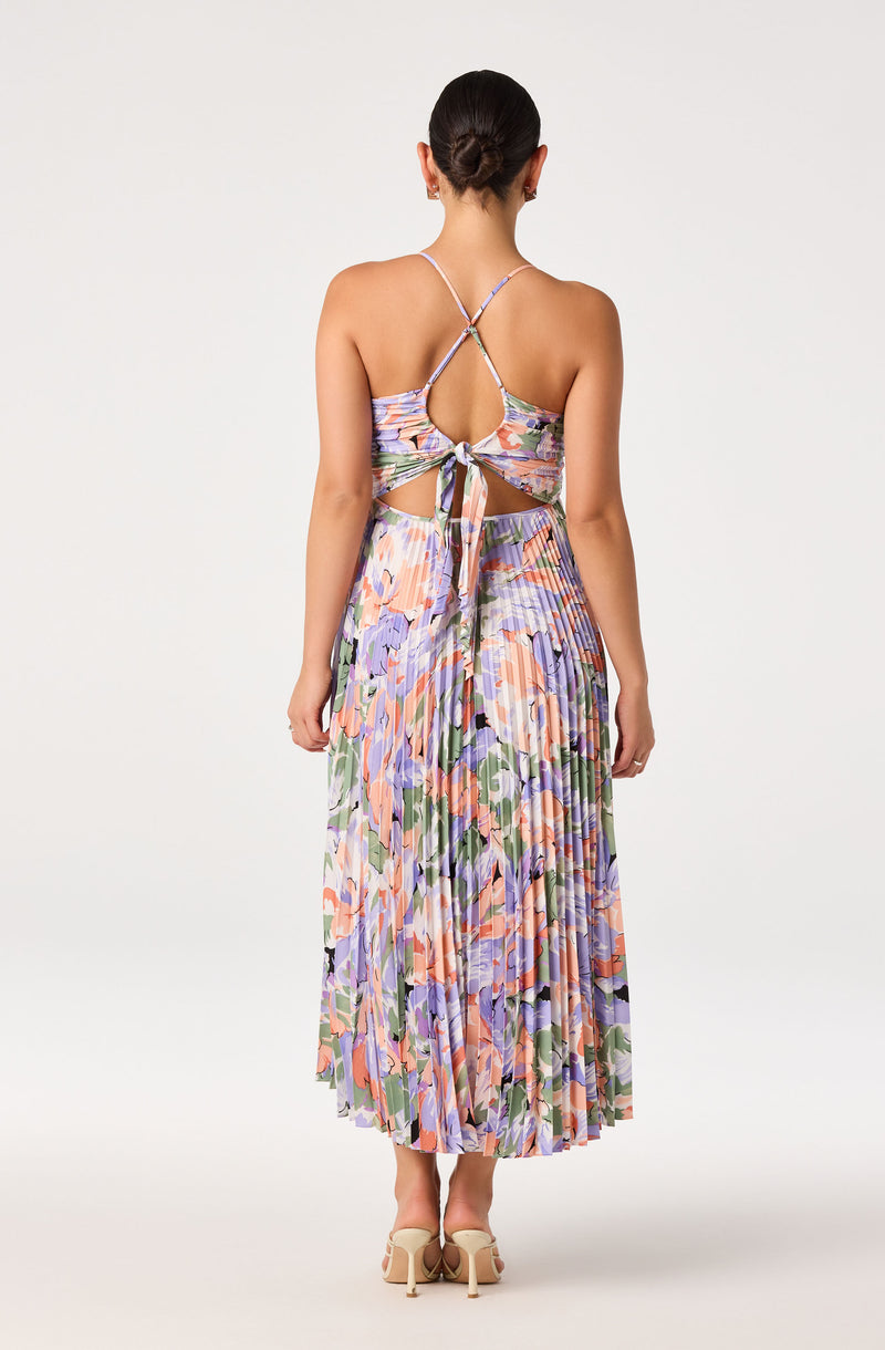 Back view of a woman wearing a multicolored floral, pleated, spaghetti strap dress with cutout and bow.