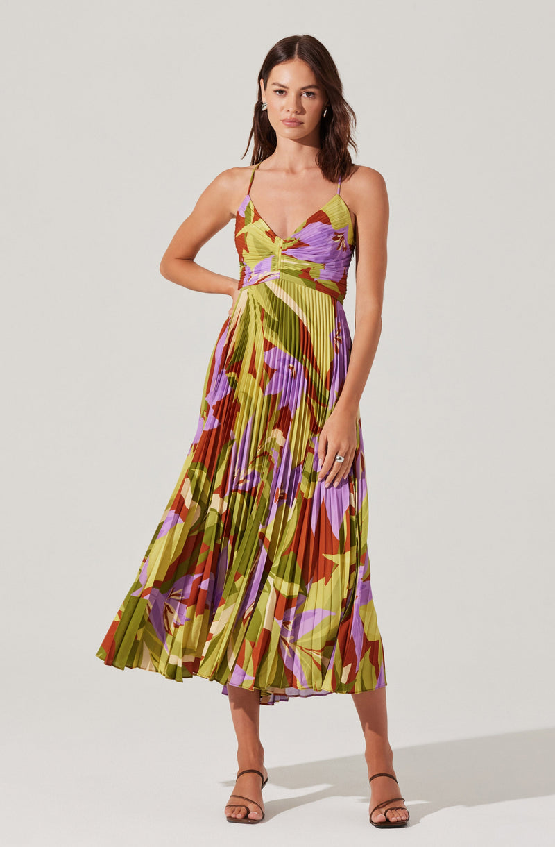 A woman is wearing a sleeveless, V-neck midi dress with spaghetti straps. The dress features a colorful, abstract floral pattern with shades of green, purple, orange, and pink, and has pleated fabric that creates a flowing silhouette. She is also wearing open-toed, ankle-strap high heel sandals.