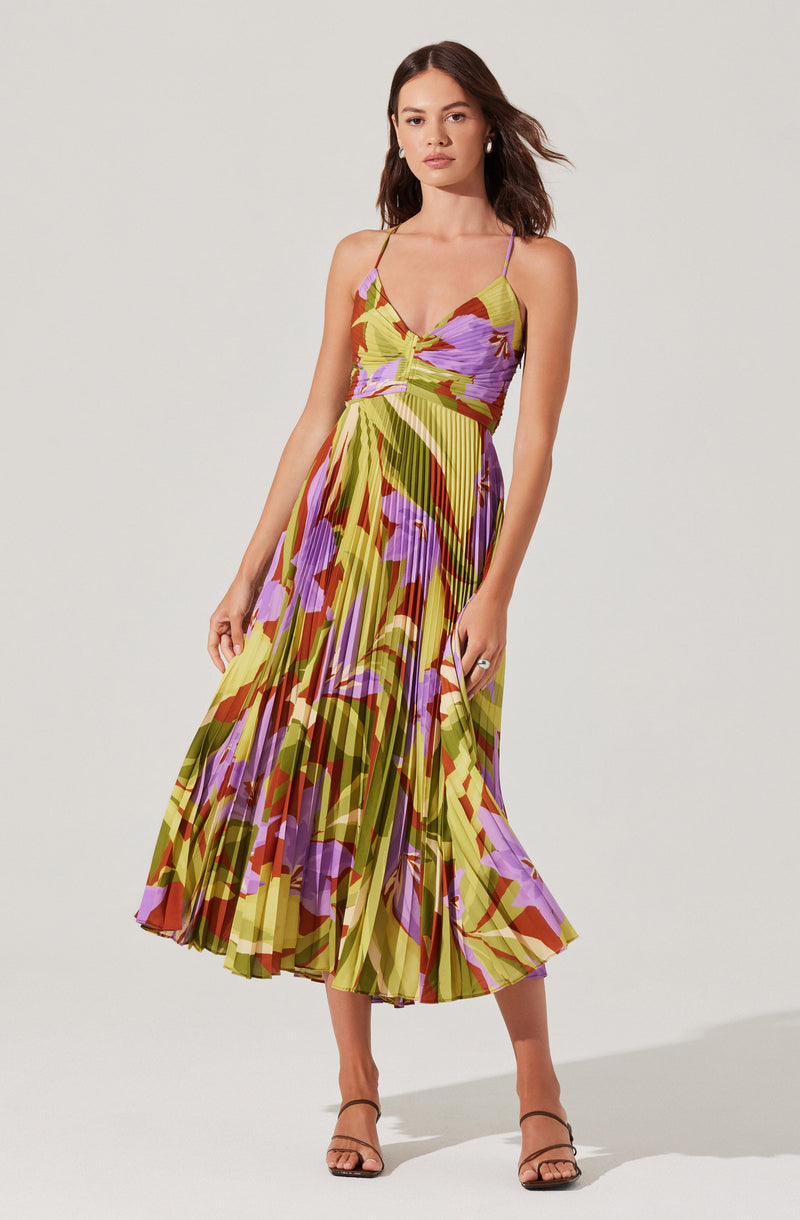 A woman is wearing a sleeveless, ankle-length dress with thin spaghetti straps. The dress features a vibrant, abstract floral pattern in shades of green, purple, yellow, and red, with a pleated skirt that adds texture and movement.