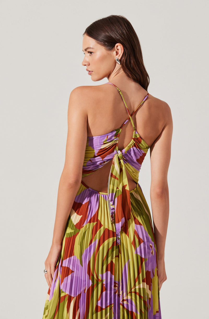 A woman is wearing a colorful, sleeveless dress with thin spaghetti straps. The dress features a vibrant, floral pattern with shades of purple, red, orange, green, and yellow. The top has a cut-out design with a tied bow at the back, creating an open-back style with fabric gathered around the tie. The skirt is pleated and flows down in a matching floral print.