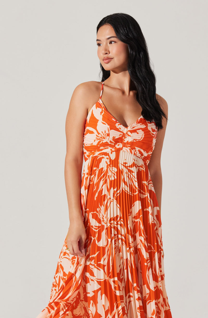 A woman is wearing a sleeveless, ankle-length dress with spaghetti straps. The dress features a bright orange background with a white floral pattern and pleats running vertically down the fabric.