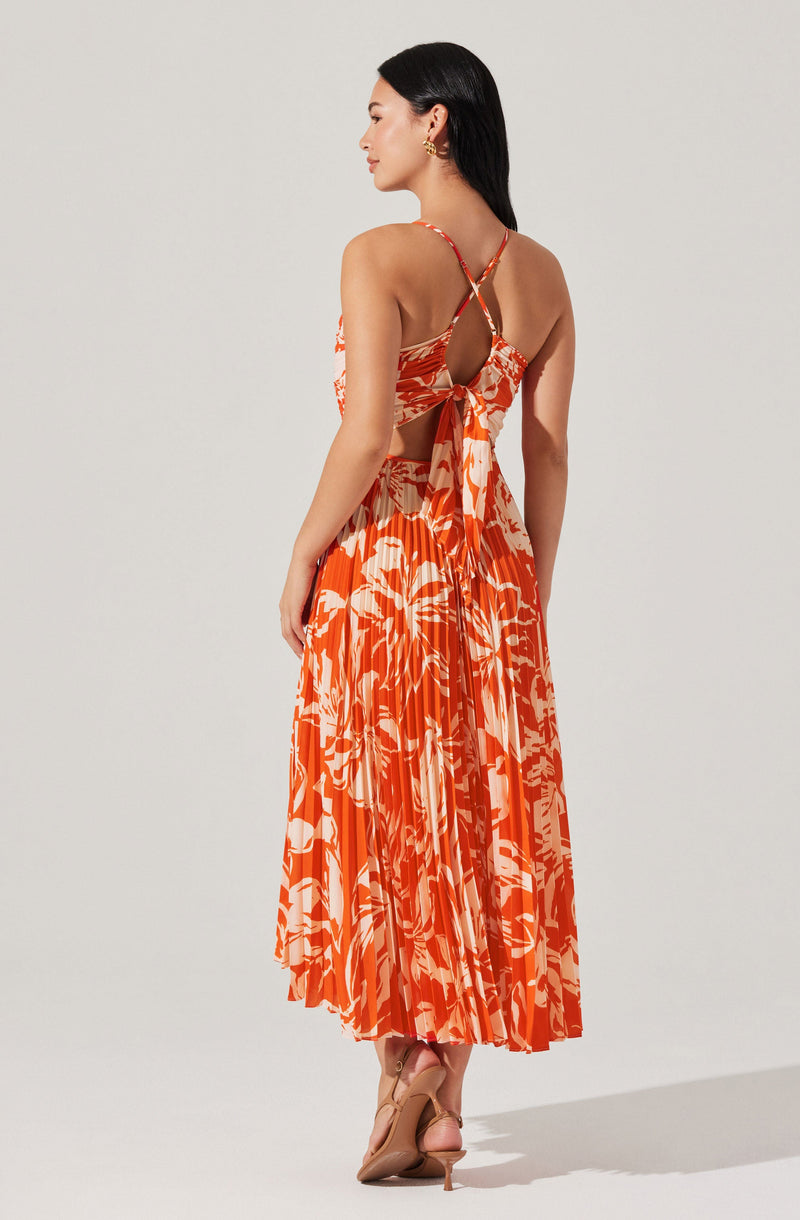 A woman is wearing a sleeveless, ankle-length dress with a vibrant orange and white abstract floral pattern. The dress features thin, cross-over spaghetti straps that tie at the back with an open back design, and a large bow detail at the waist. She is also wearing nude high-heeled shoes.