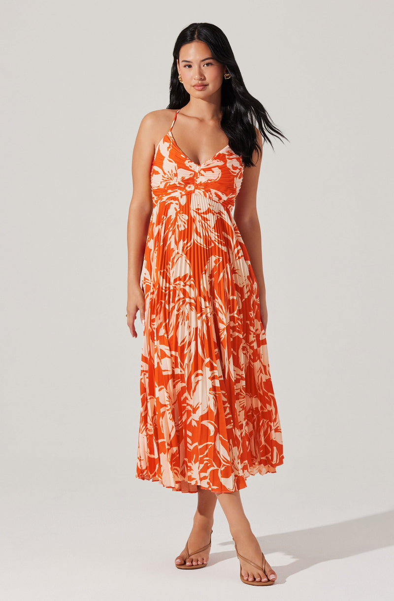 A woman is wearing a sleeveless, orange maxi dress with a white floral and leaf pattern. The dress has thin spaghetti straps, a V-neckline, and an empire waistline. The skirt is flowing and pleated, reaching down to her ankles. She is also wearing clear, strappy high-heeled sandals.