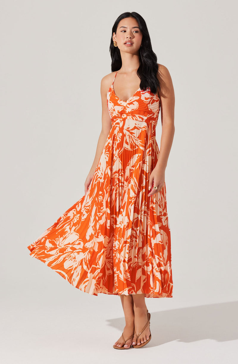 A woman is wearing a sleeveless, ankle-length dress with thin spaghetti straps. The dress features a vibrant floral pattern in shades of orange and white, with a fitted bodice and a flowing, pleated skirt. She is also wearing nude-colored heels and gold earrings.