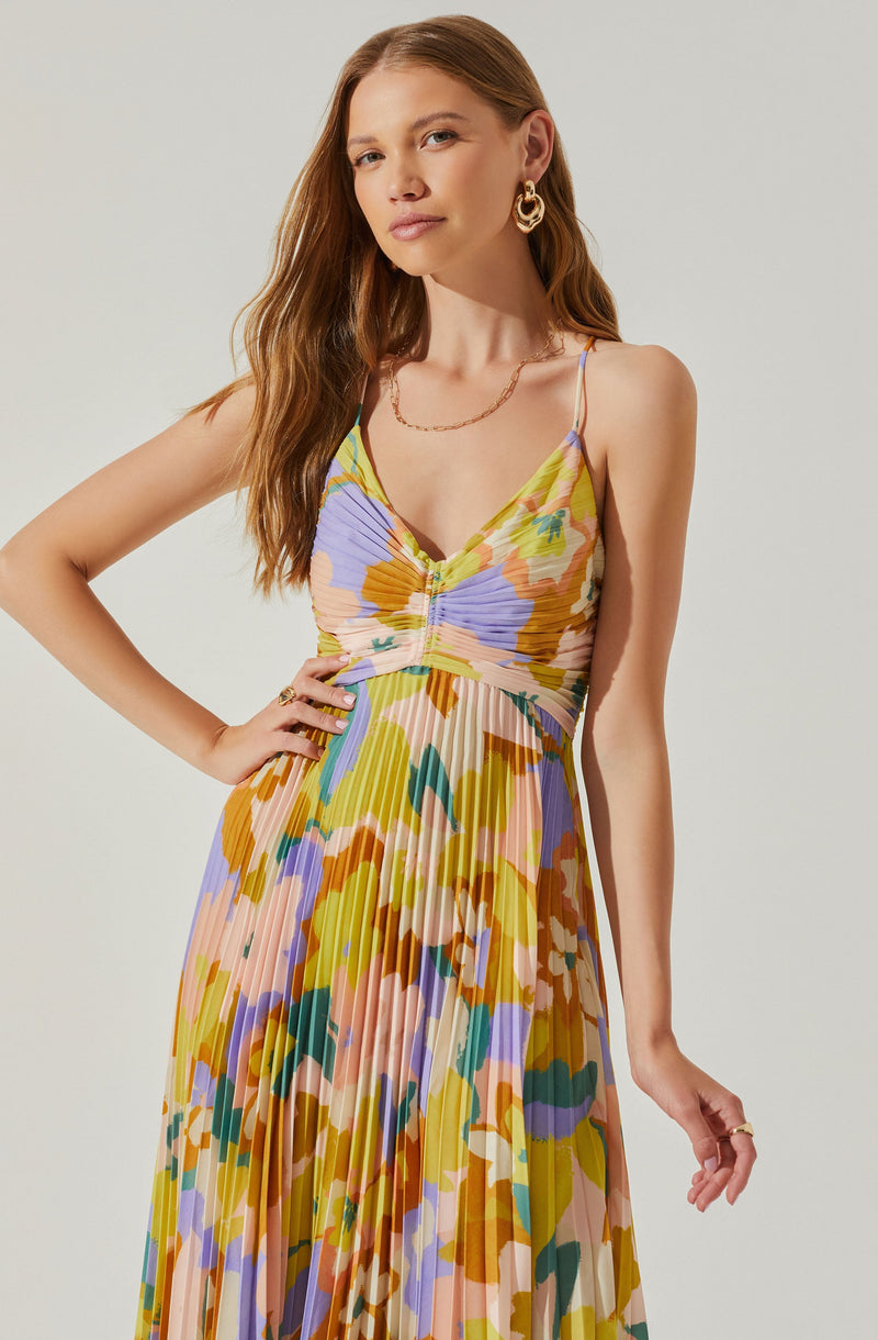A woman is wearing a sleeveless, v-neck maxi dress with thin spaghetti straps. The dress features a colorful, abstract floral pattern with shades of yellow, purple, pink, orange, green, and cream, and includes pleats that add texture and movement to the fabric. She accessorizes with gold hoop earrings, a delicate gold chain necklace, and a ring on her right hand.