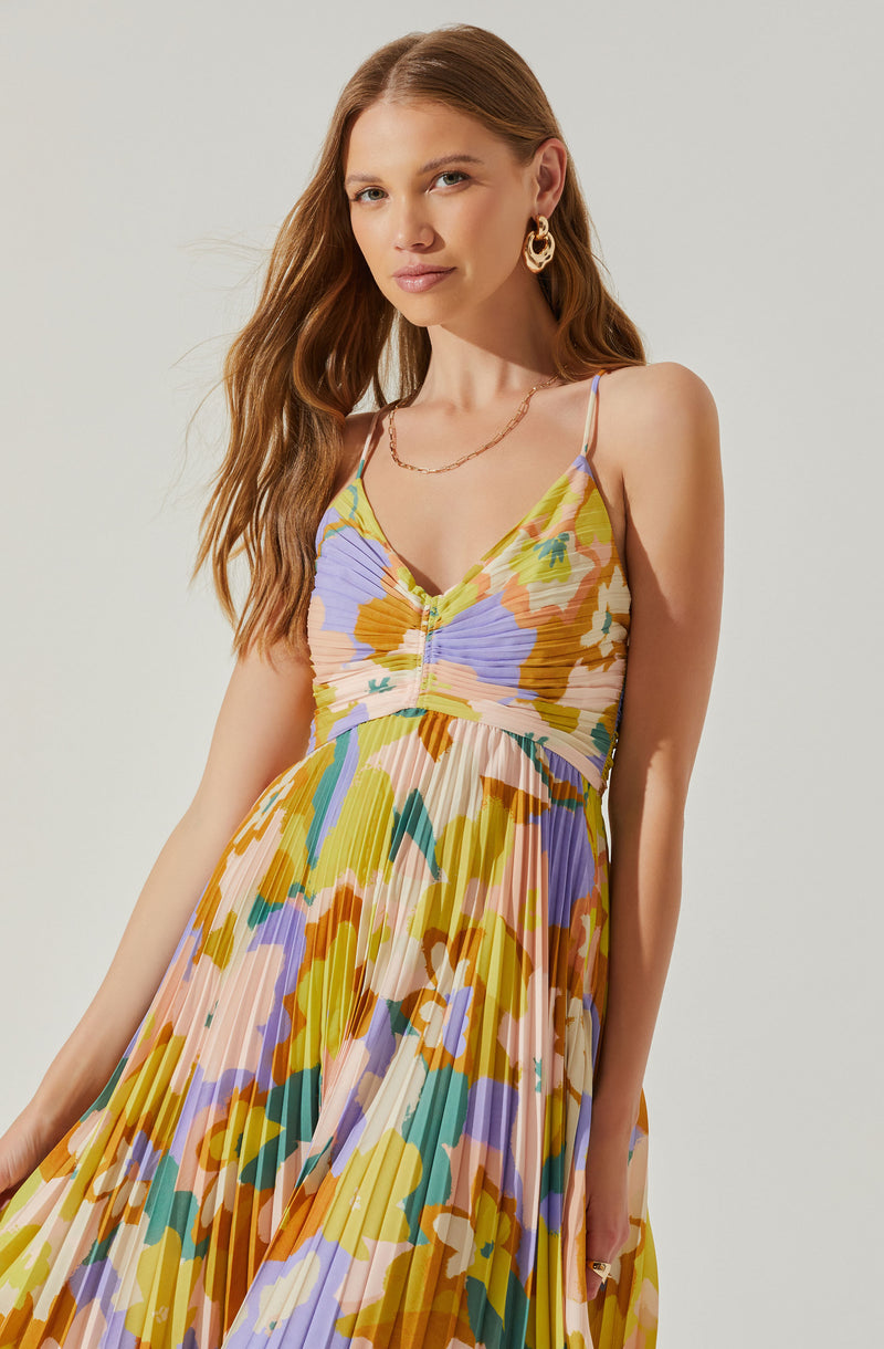 A woman is wearing a sleeveless, V-neck maxi dress with thin spaghetti straps. The dress features a colorful, floral abstract pattern with shades of pastel yellow, purple, green, orange, and cream. The fabric is pleated, creating a textured, flowing appearance.