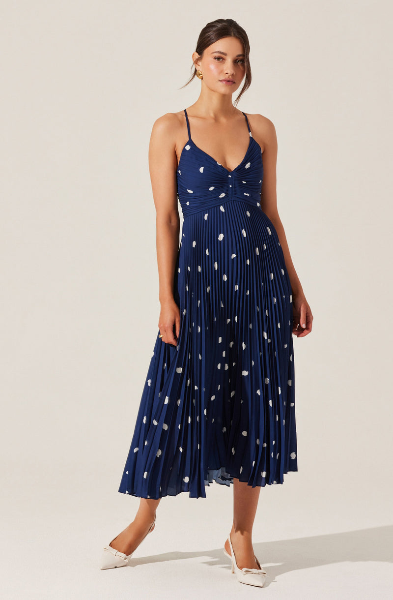 A woman is wearing a navy blue pleated dress with white polka dots and spaghetti straps. The dress has a fitted bodice with ruching details. She is also wearing white heels and gold earrings.