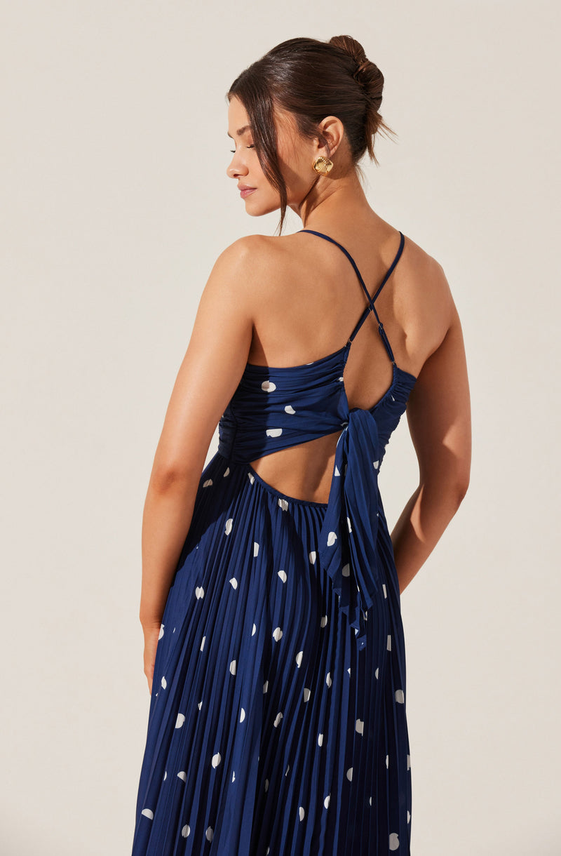 A woman is wearing a navy blue dress with white polka dots. The dress features thin spaghetti straps that cross at the back, with a tied bow detail just below the back. The skirt has a pleated design. She is accessorized with large, gold, shell-shaped earrings.
