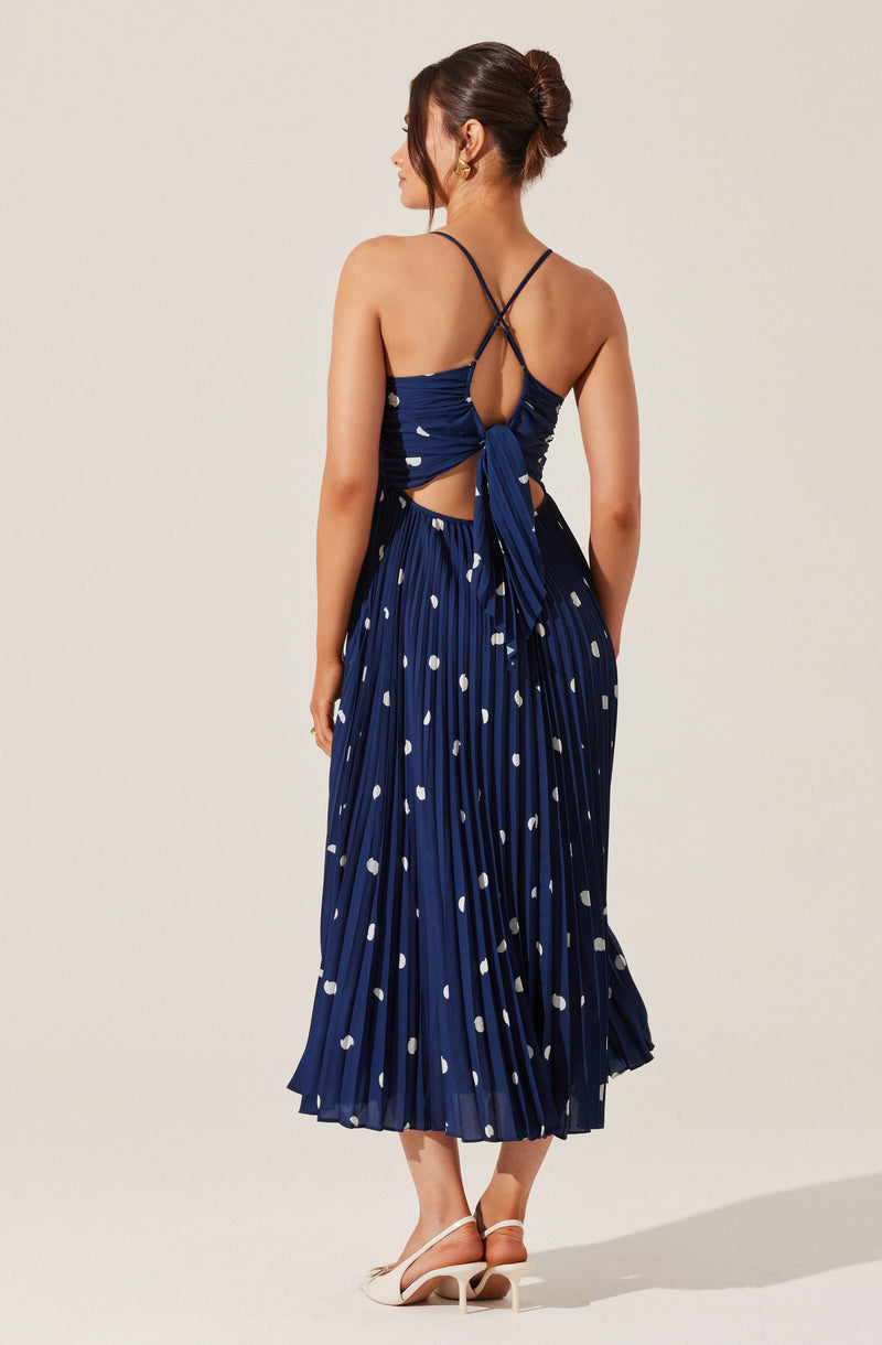 The dress is a navy blue, sleeveless, pleated maxi dress with white polka dots. It features spaghetti straps that cross at the back and tie into a bow, creating an open-back design with a keyhole opening at the upper back. The hemline reaches just above the ankles.