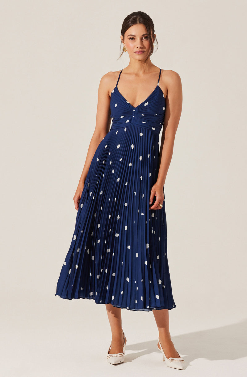 A woman is wearing a navy blue, sleeveless, spaghetti strap dress with a fitted bodice and a flared, pleated skirt. The dress features a pattern of irregular white dots throughout. She is also wearing white high-heeled shoes with bows at the front.
