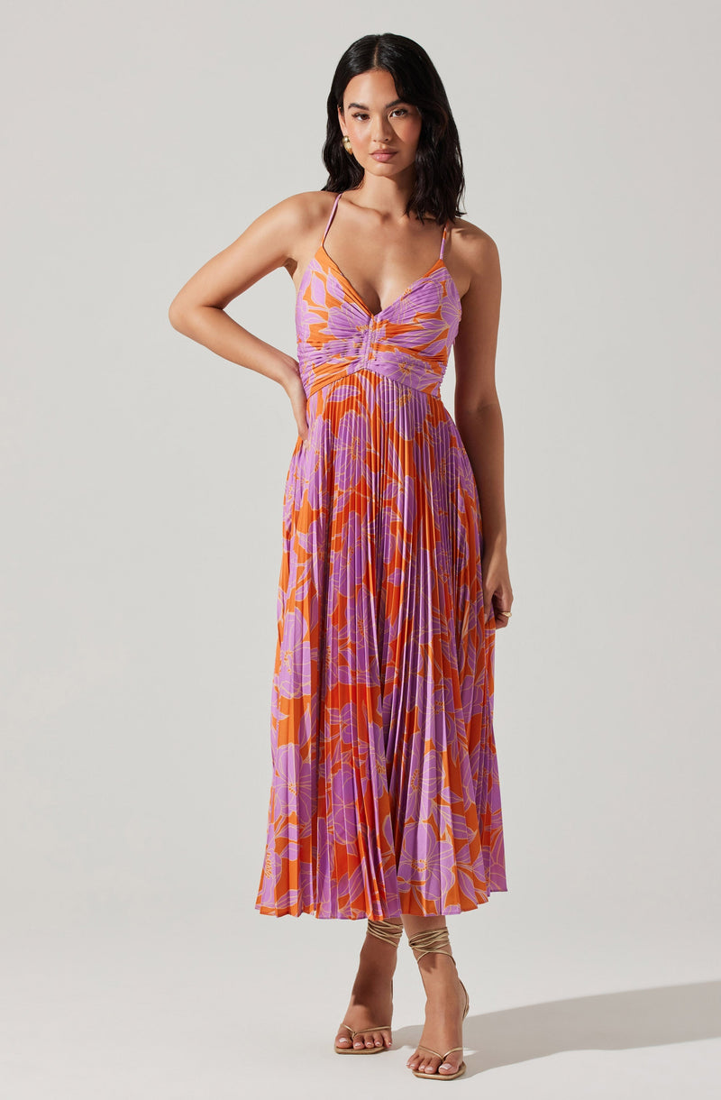 A woman is wearing a sleeveless, floor-length dress with a vibrant floral pattern in shades of orange, purple, and pink. The dress features a ruched bodice and thin spaghetti straps. She pairs it with open-toed, nude heels that have ankle straps.