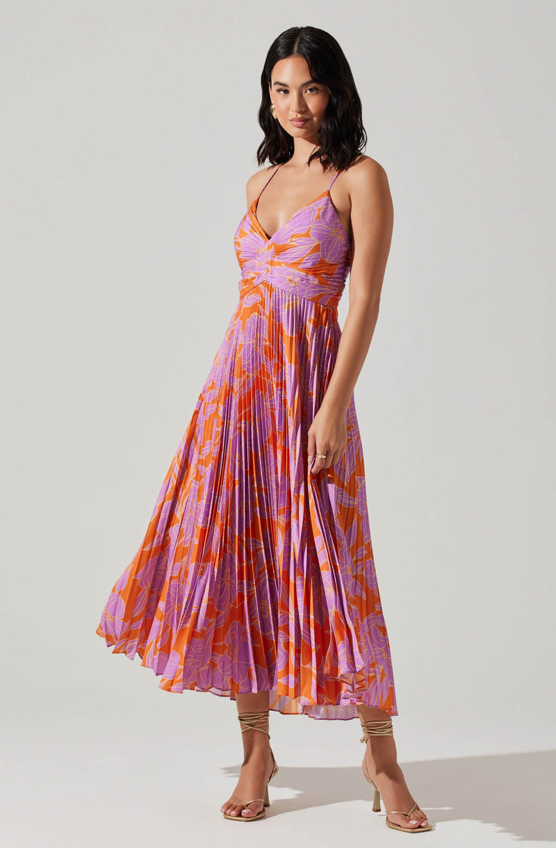 A woman is wearing a sleeveless, floor-length dress with thin spaghetti straps. The dress features a colorful abstract floral pattern in shades of orange, purple, and pink. She pairs the dress with nude high-heeled sandals that have ankle straps.
