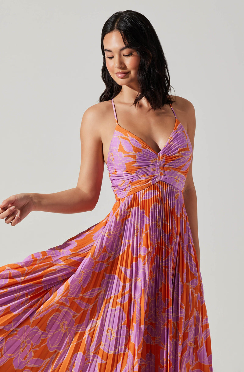A woman is wearing a sleeveless, flowy dress with thin spaghetti straps. The dress features a vibrant floral pattern with large purple flowers and orange background. The fabric appears lightweight and has a pleated design, creating a soft, flowing effect. The top of the dress has a ruched detail at the bust, adding some texture to the garment.