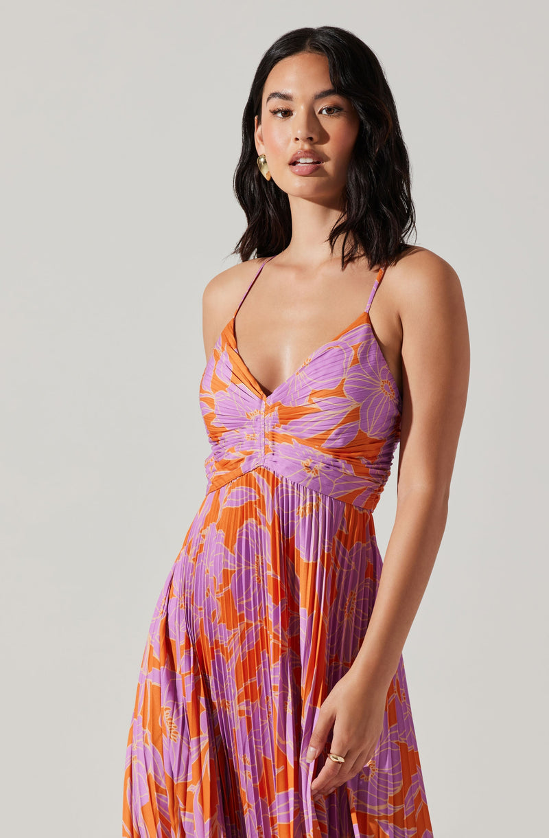 A woman wearing a sleeveless, flowy dress with thin spaghetti straps. The dress features a vibrant floral pattern in shades of purple, orange, and pink, with a gathered bust detail and a pleated skirt that falls gracefully.