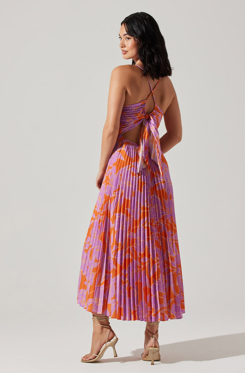 A woman is wearing a colorful dress with a vibrant, abstract floral pattern in purple, orange, and pink. The dress has thin spaghetti straps and features a large bow tied at the back with a cutout design. The skirt part of the dress is pleated and reaches mid-calf. She is also wearing nude high-heeled sandals with ankle straps.
