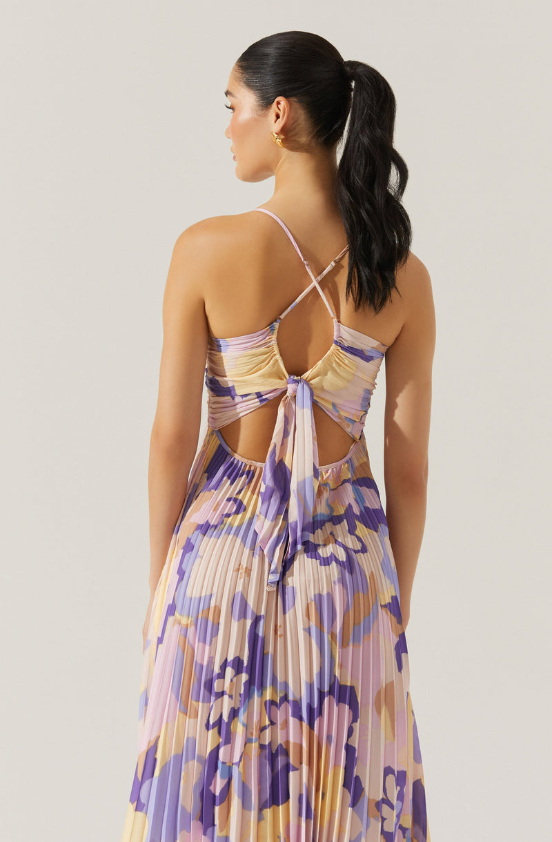 A woman is wearing a sleeveless dress with thin spaghetti straps that crisscross at the back. The dress features a fitted bodice with a bow tied at the lower back, and a flowing skirt with an abstract floral pattern in shades of purple, beige, cream, and light pink, with pleated fabric.