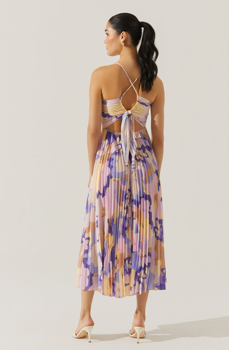 A woman is wearing a sleeveless, pleated dress with a multicolored floral pattern in shades of purple, pink, yellow, and beige. The dress features thin spaghetti straps, a cutout back with a tie at the waist, and a flowing midi length. She is wearing beige high-heeled shoes. She has long, dark hair tied back in a high ponytail and is accessorized with gold earrings.