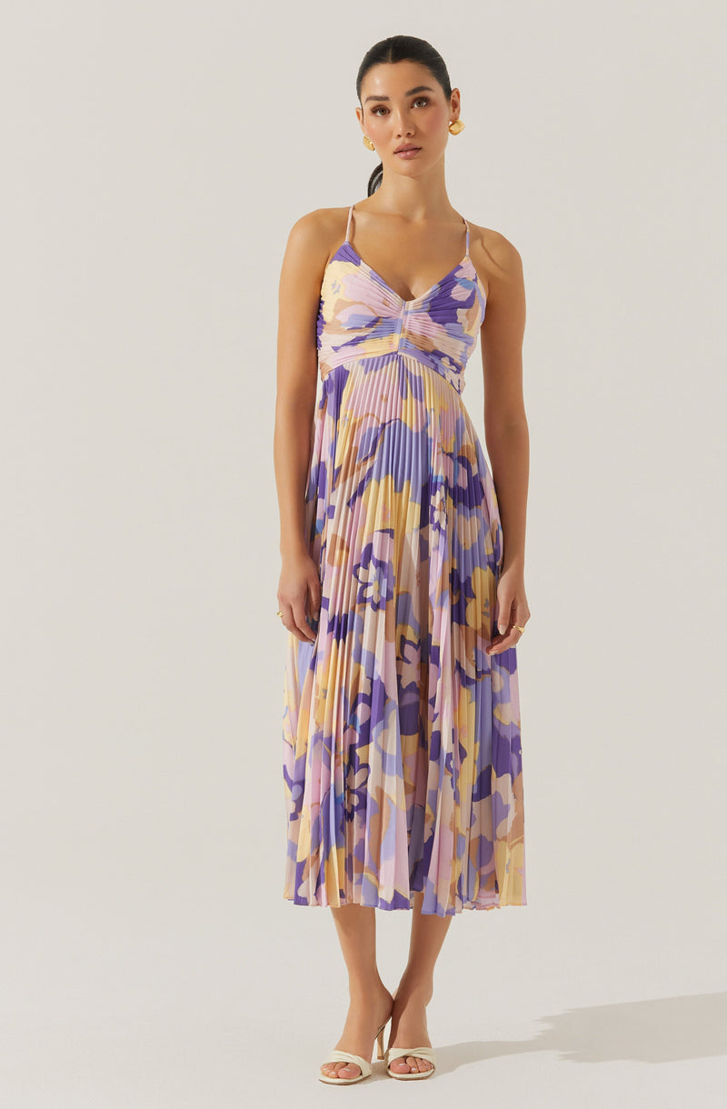 A woman is wearing a sleeveless, V-neck, ankle-length dress with thin spaghetti straps. The dress features a colorful abstract floral pattern in shades of purple, pink, yellow, and beige, with a pleated skirt and ruched bodice. She is accessorized with gold hoop earrings and appears to be wearing open-toe sandals.
