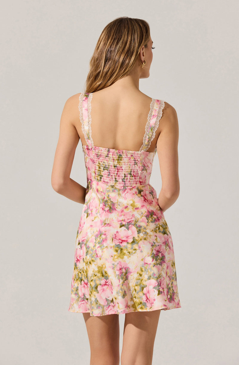 A woman is wearing a sleeveless dress with wide straps decorated with pink and white lace trim. The dress features a smocked bodice and a floral pattern with pink, green, and cream colors on a light background. The skirt of the dress is flared and short.