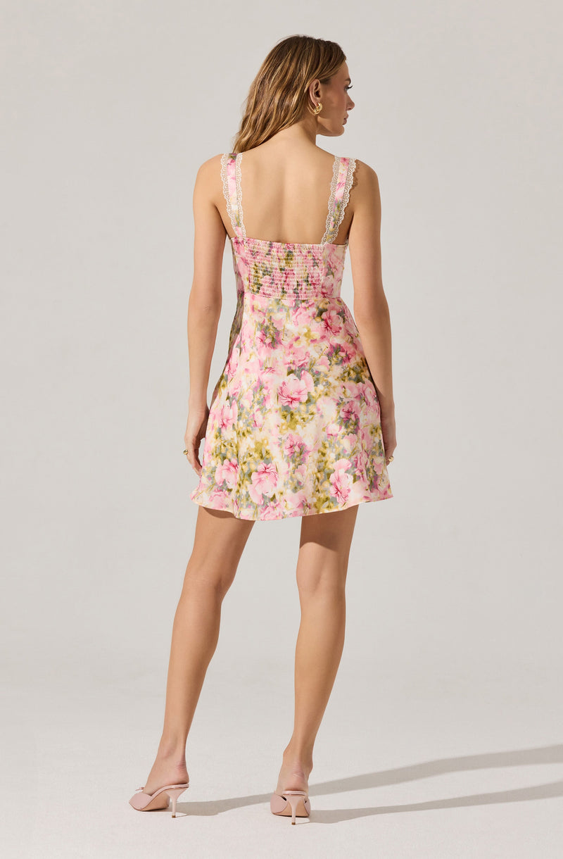 A woman is wearing a sleeveless, ruffled-strap dress with a fitted, shirred bodice and a short, flared skirt. The dress features a floral pattern with pink, green, and white tones.